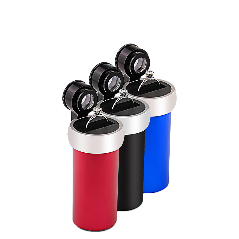 Three variants of the JYBS diamond inscription loupe in red, black, and blue finishes, each holding a silver engagement ring on its platform stage. Three variants of the JYBS diamond inscription loupe in red, black, and blue finishes, each holding a silver engagement ring on its platform stage.