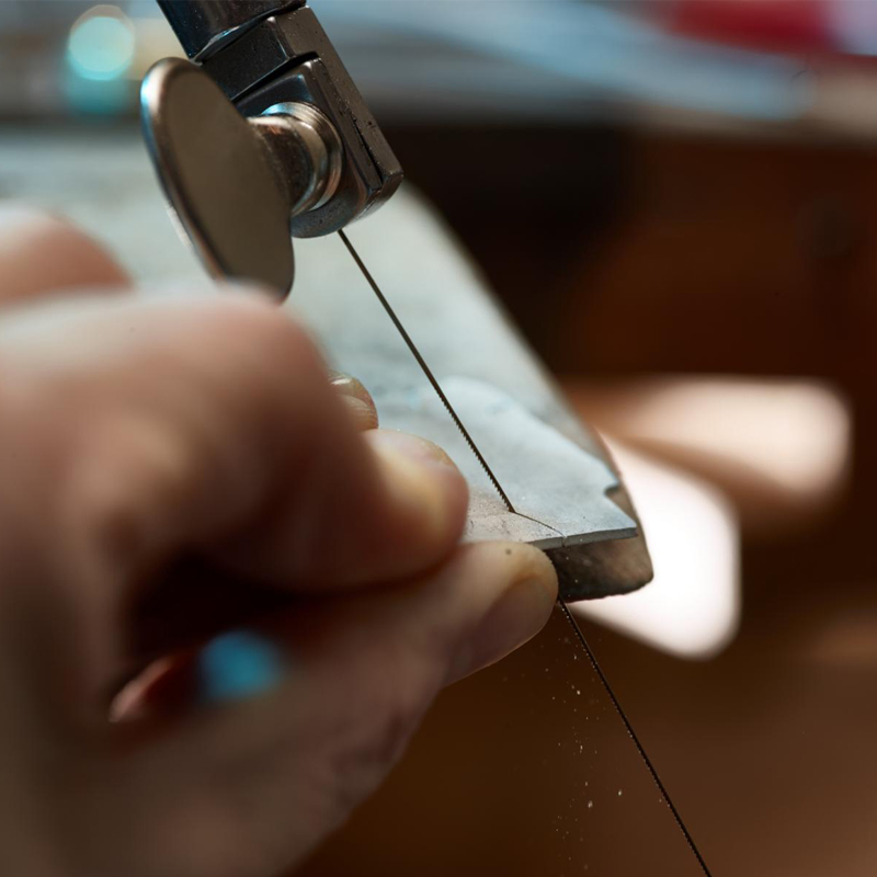Close-up of a hand using a jeweler's saw frame and a fine saw blade to cut a metal sheet on a bench pin. Close-up of a hand using a jeweler's saw frame and a fine saw blade to cut a metal sheet on a bench pin.