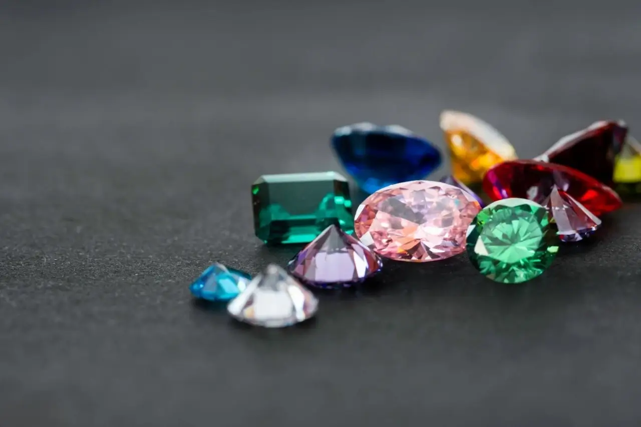 A variety of colorful faceted gemstones are scattered on a dark surface, with a large pink oval gem in the center