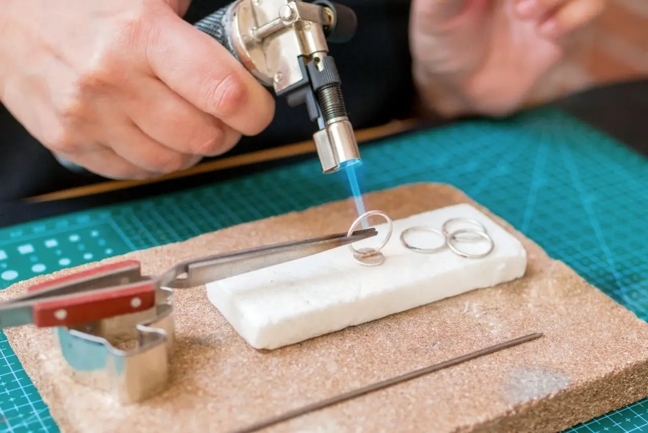 a close up of a jeweler using a micro torch with a blue flame to heat a silver ring held by tweezers