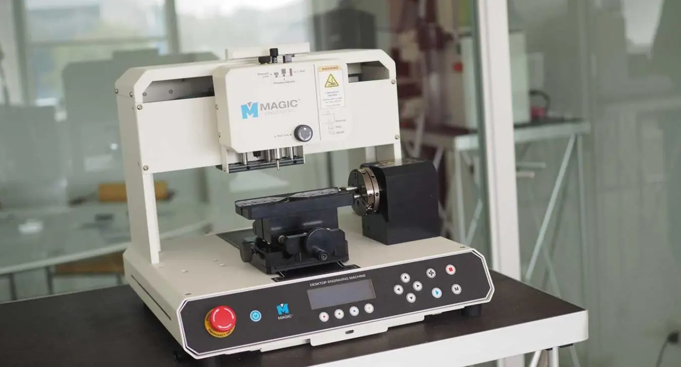 A white desktop engraving machine, model MAGIC, with a rotary attachment and control panel on a black table.