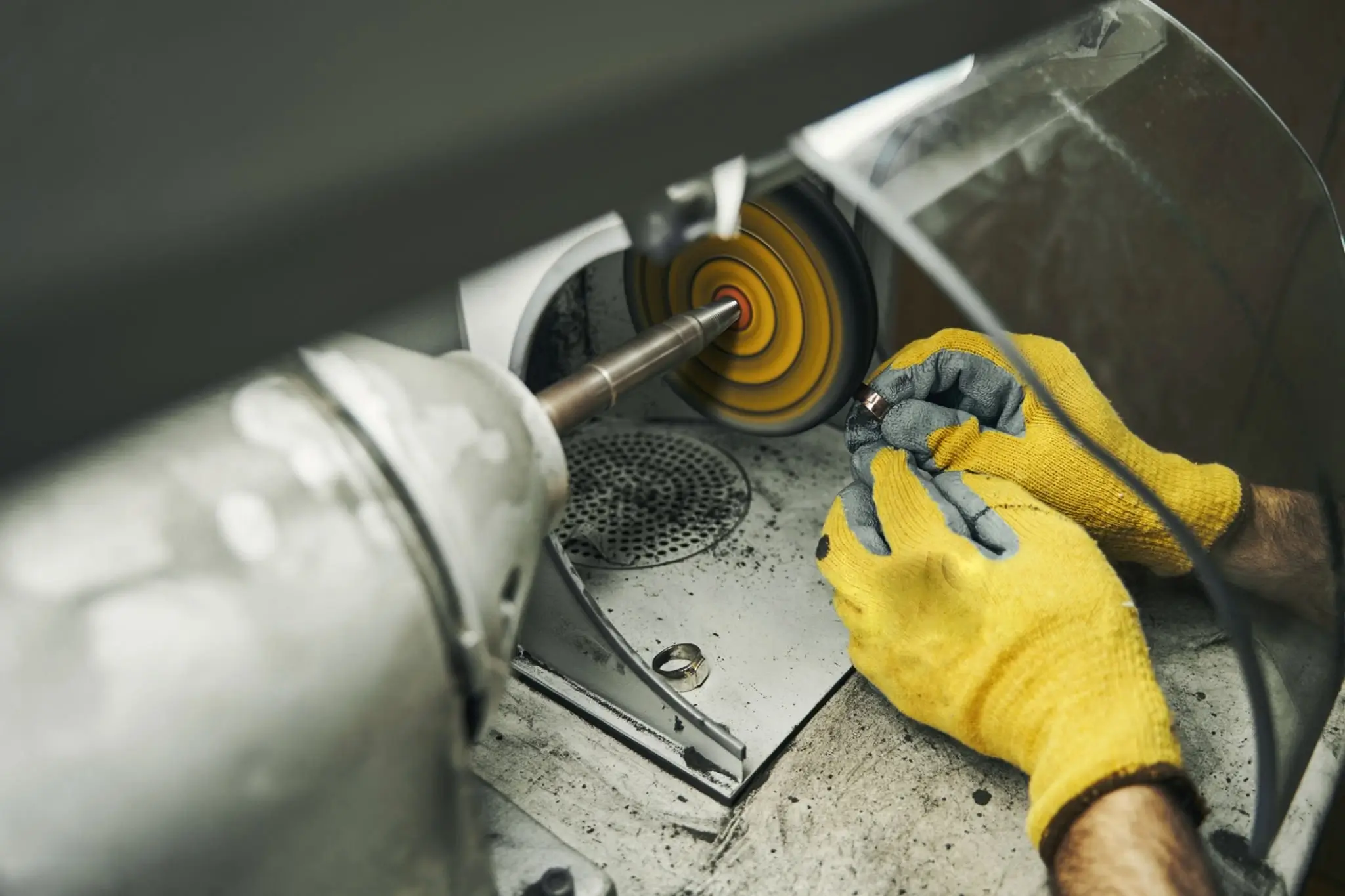 Jeweler in yellow gloves polishing a piece of jewelry using a rotary polishing machine with a felt wheel.