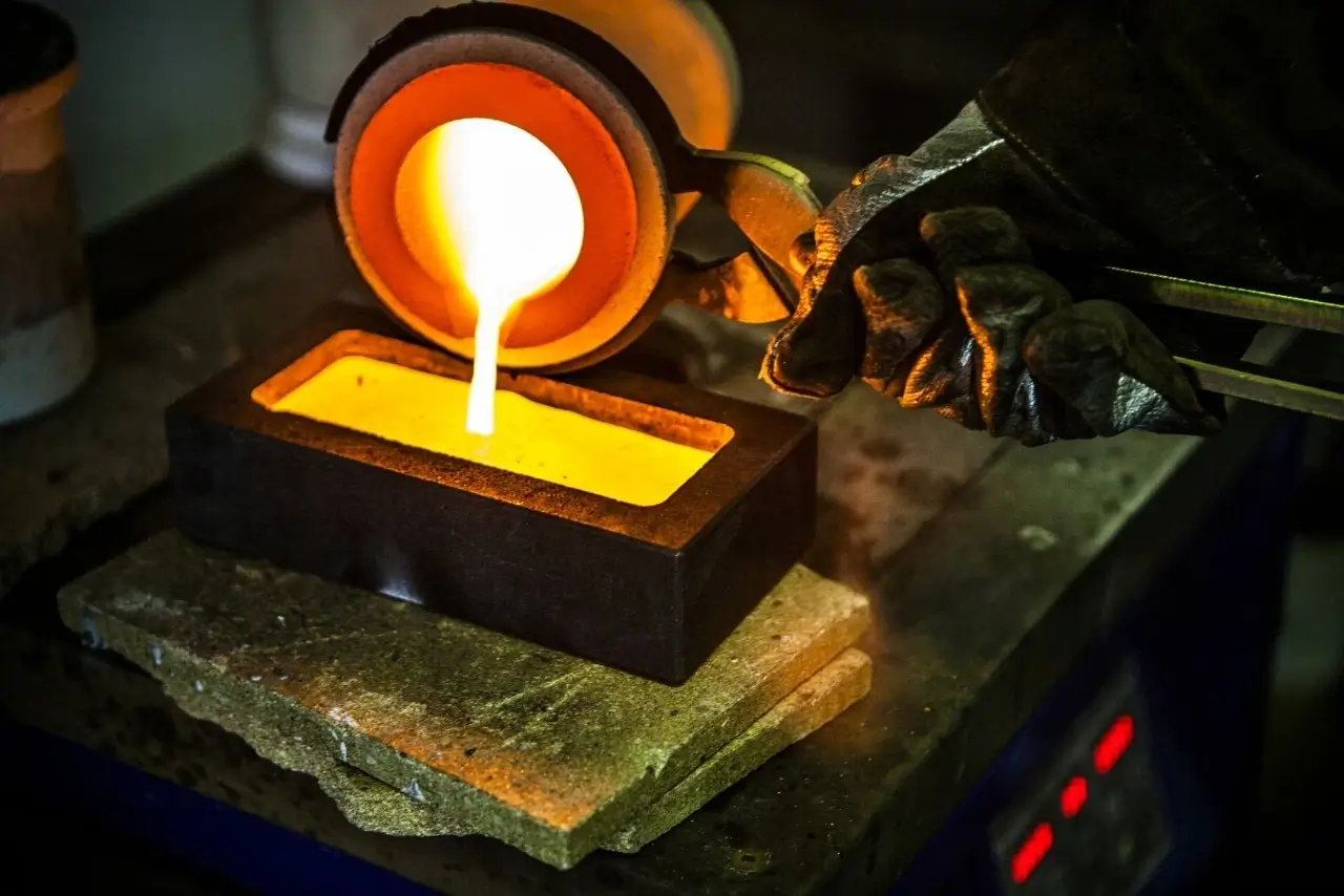 a gloved hand holds a crucible pouring molten gold into a rectangular mold glowing with intense heat