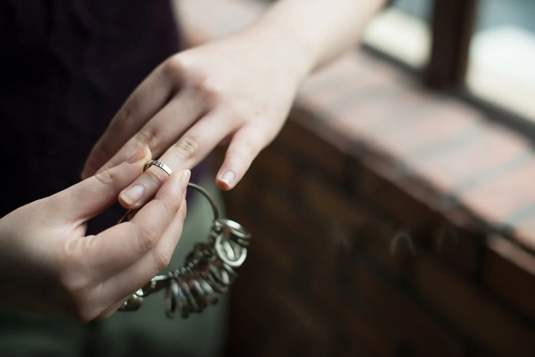 close up of a persons hands holding a set of ring sizers and trying a silver band on their finger near a window
