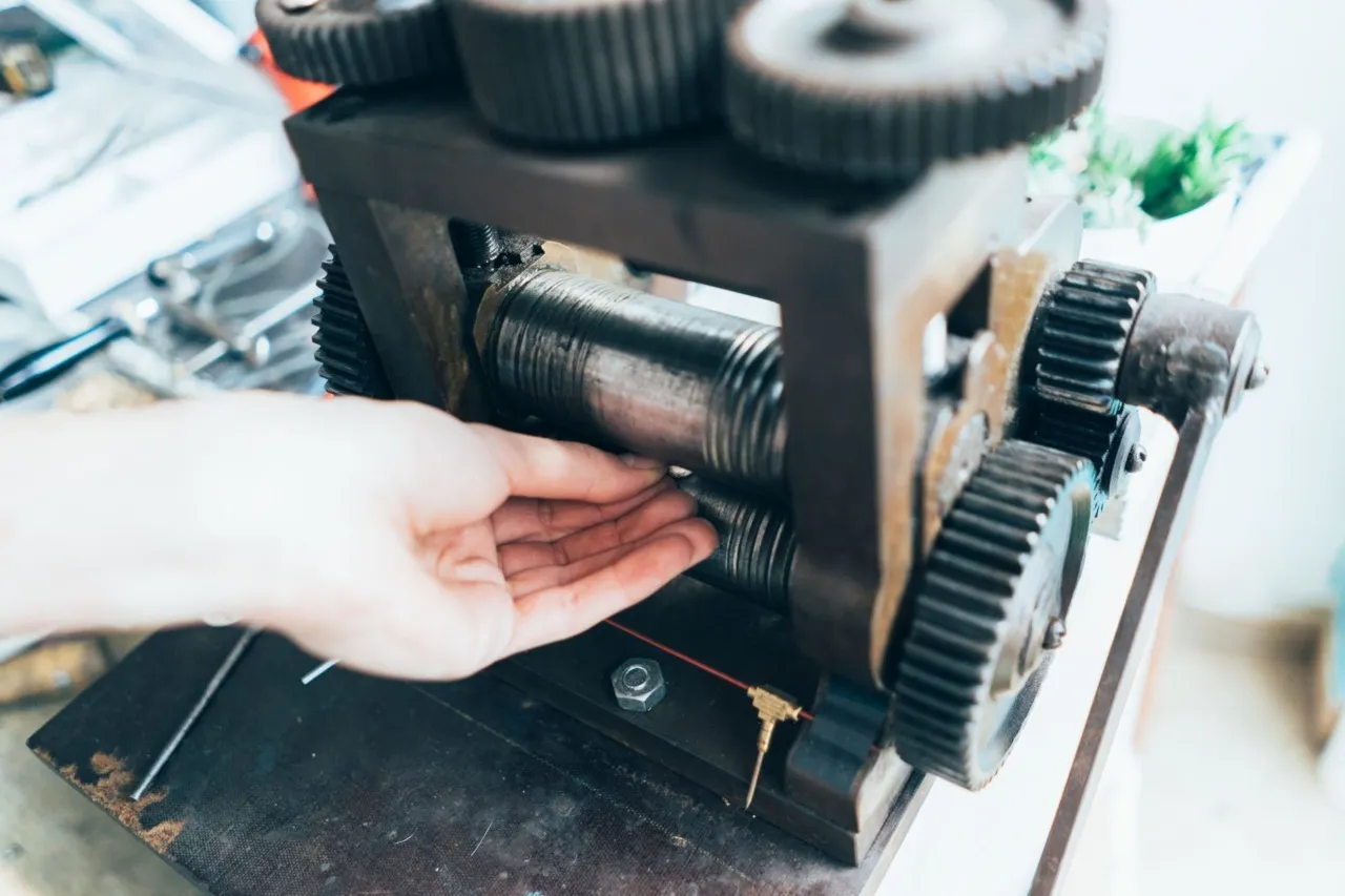 Close-up of a hand feeding a small piece of metal between rollers on a manual rolling mill for recycling.