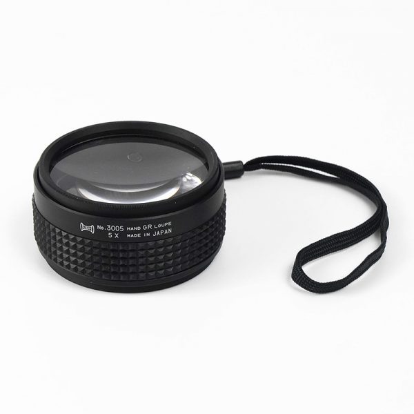 A 5X magnification Hand Grip Loupe with its black lanyard resting on a white surface