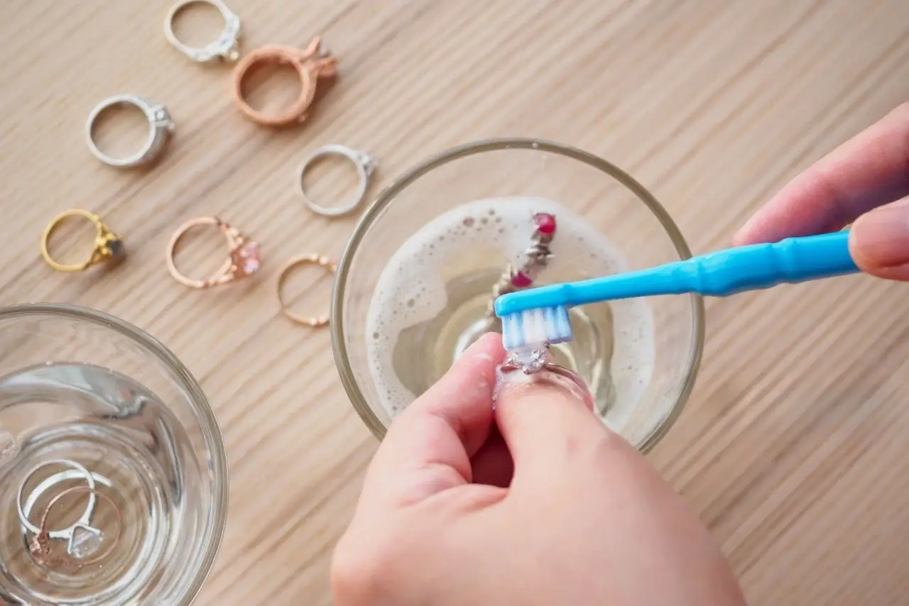 a hand using a toothbrush to clean a ring in a glass of soapy water. Other clean rings and another glass of water are nearby