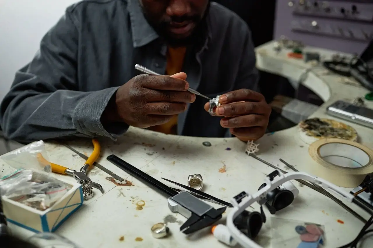 A male jeweler in a gray shirt is carefully crafting a piece of jewelry with a tool at his workbench, surrounded by various tools and materials