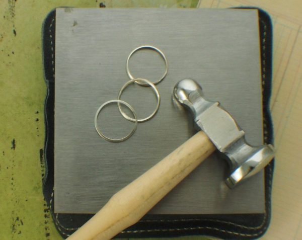 A square black jeweler's sandbag with a metal dapping block, hammer, and three silver rings on top