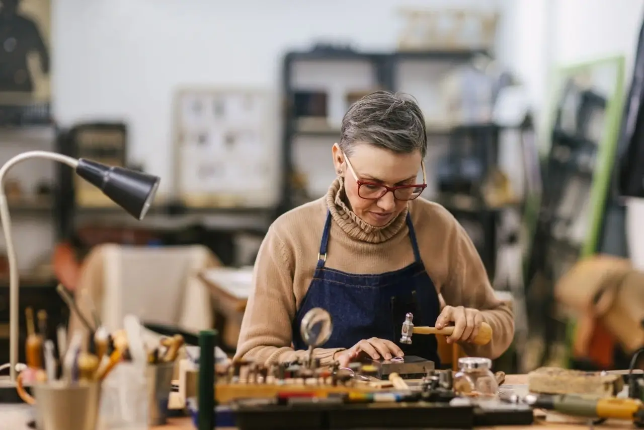 A woman with glasses and an apron using a hammer and bench block to make jewelry at a cluttered workbench