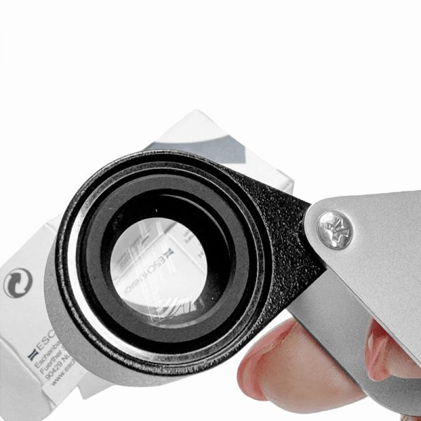 A hand holding an Eschenbach magnifier up close to its product box for inspection.