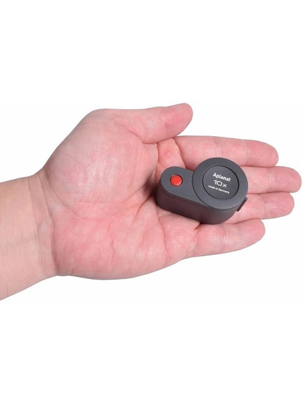 A hand holds the Eschenbach Aplanat 10x magnifier, showing its compact size.