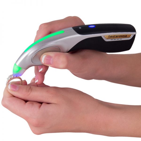 A person uses the GemOro Testerossa to test a diamond ring, showing a green light for a positive result.