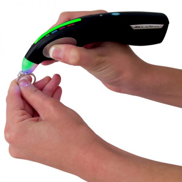 A person uses the GemOro UltraTester 3+ to test an engagement ring, with a green light indicating a diamond.