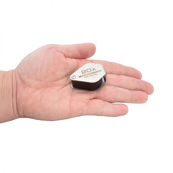 An Eschenbach 20x folding magnifier held in the palm of a hand for a sense of scale.