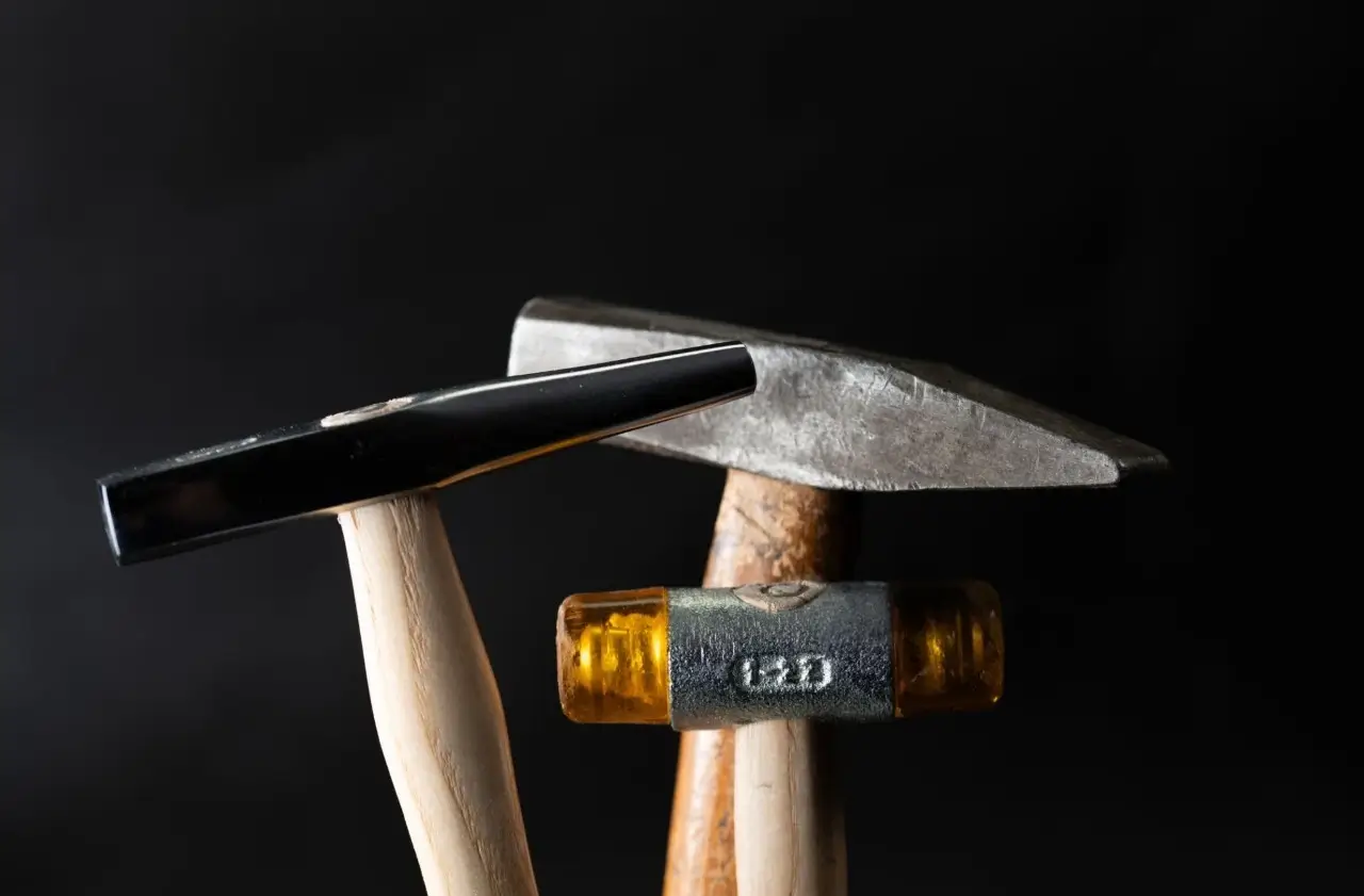 Close-up of three jewelry hammers, including a chasing hammer, a planishing hammer, and a small yellow rubber mallet, on a dark background.